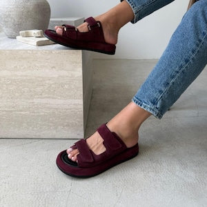 May include: Burgundy sandals with two adjustable straps. The sandals have a thick sole and a black footbed. The straps are secured with metal buckles. The sandals are worn with blue jeans.