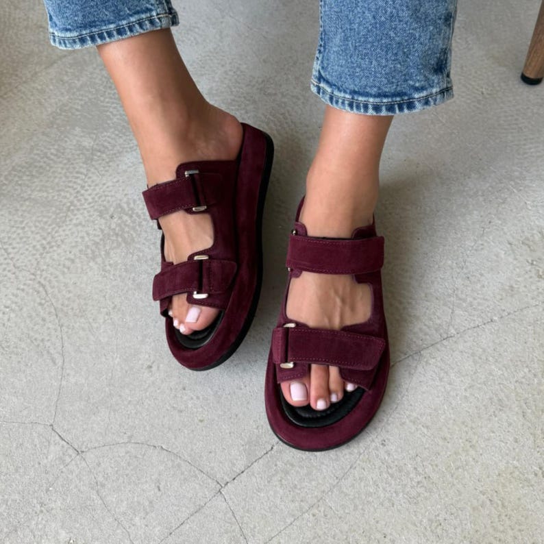 May include: Burgundy suede sandals with two adjustable straps and silver buckles. The sandals have a thick sole and are shown on a light gray surface. The footwear is a stylish choice for casual wear.