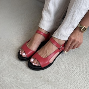 Puede incluir: Sandalias de cuero rojas con suela negra y correa en el tobillo. Las sandalias tienen cierre de hebilla y se llevan con pantalones claros. Las sandalias son de color rojo brillante.