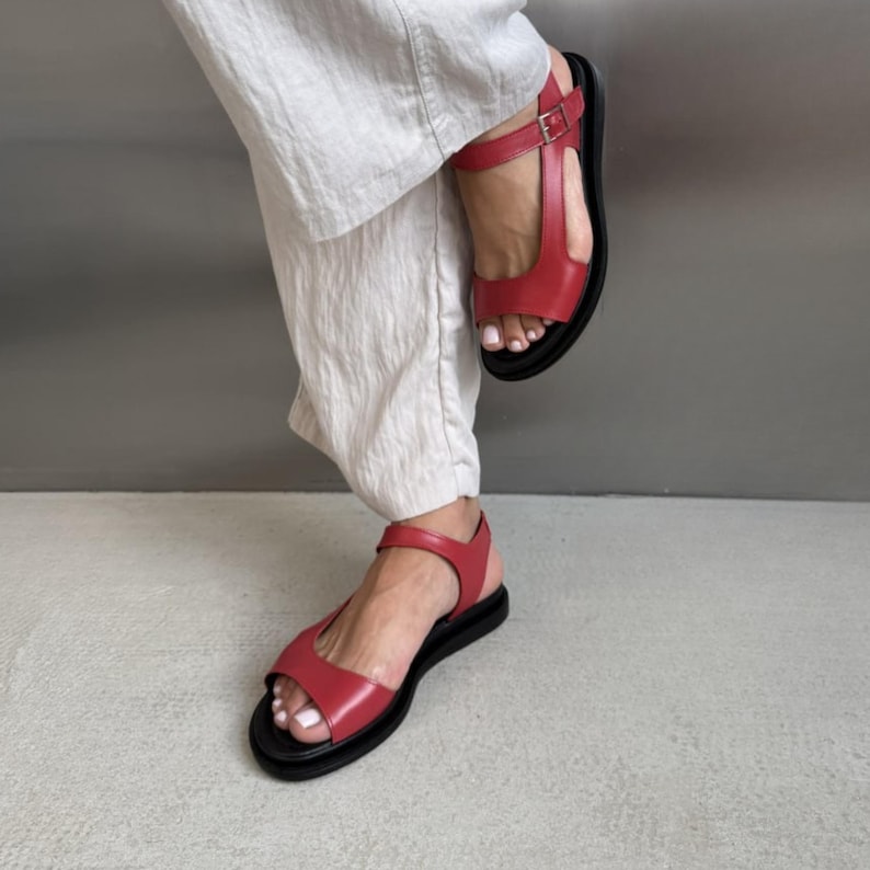 Puede incluir: Sandalias de cuero rojo con suela negra y cierre de hebilla. Las sandalias tienen un dise&ntilde;o de tira en T y suela plana. Las sandalias se muestran con pantalones blancos.