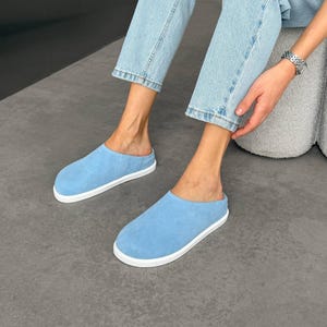 Può includere: Un paio di scarpe slip-on in camoscio azzurro con punta arrotondata e una spessa suola in gomma bianca. Le scarpe presentano una struttura semplice senza tallone con una finitura opaca liscia sulla tomaia e un profilo di colore solido.