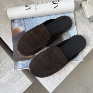 Puede incluir: Un par de mocasines sin cordones de ante marrón con diseño de mocasín de centavo. Los zapatos tienen una parte superior marrón oscuro y una suela negra. Los zapatos se exhiben en una revista.
