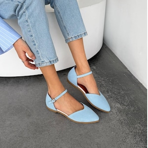May include: Light blue flats with ankle straps. These shoes feature a pointed toe design and a small buckle closure on the ankle strap. The soles are a light brown color, and the interior is white. The flats are made of a soft, suede-like material.