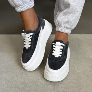 May include: A pair of platform sneakers with a dark gray suede upper and white soles. The sneakers have white laces and a rounded toe. The shoes are on a gray surface.