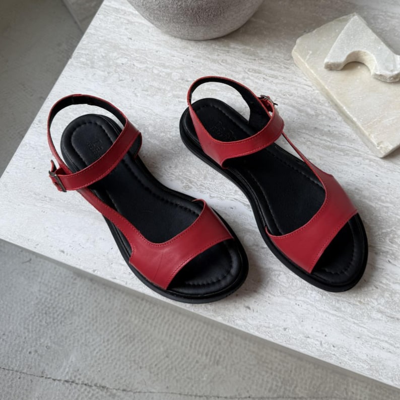 Puede incluir: Un par de sandalias de cuero rojo con suelas negras. Las sandalias presentan un dise&ntilde;o de correa en T y correas de tobillo ajustables con hebillas. Las sandalias de punta abierta se exhiben sobre una superficie clara.