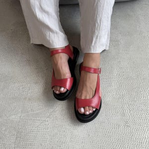 Puede incluir: Sandalias de cuero rojo con suela negra y correa en el tobillo. Las sandalias tienen cierre de hebilla y punta redondeada. La imagen muestra las sandalias desde arriba.
