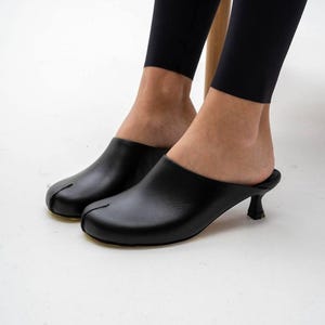 Puede incluir: Mules de cuero negro con diseño de puntera dividida y tacón pequeño y curvado. Los zapatos tienen un acabado liso y brillante y una estética minimalista. Las mules son de estilo sin cordones.