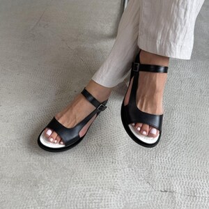 Puede incluir: Sandalias de cuero negro con suela blanca y correa en el tobillo. Las sandalias tienen cierre de hebilla y un dise&ntilde;o minimalista. El calzado se muestra sobre una superficie gris claro.