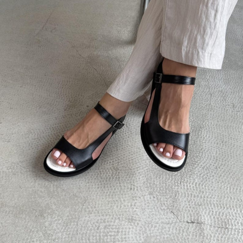 Puede incluir: Sandalias de cuero negro con suela blanca y correa en el tobillo. Las sandalias tienen cierre de hebilla y un dise&ntilde;o minimalista. El calzado se muestra sobre una superficie gris claro.