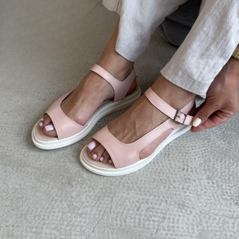 Puede incluir: Sandalias rosa p&aacute;lido con suelas blancas y correas en el tobillo. El dise&ntilde;o de punta abierta presenta una correa en la parte superior del pie y un cierre de hebilla. Las sandalias tienen un estilo moderno y minimalista.