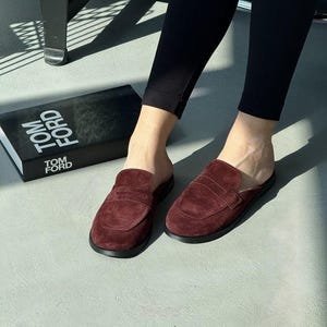 Puede incluir: Mocasines sin cordones de ante burdeos con suela negra. Los zapatos se muestran sobre una superficie gris claro, con un libro negro que dice "TOM FORD" en texto blanco.