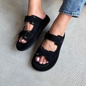 May include: Black suede sandals with two adjustable straps and silver-tone buckles. The sandals have a thick sole and are worn on a light gray surface. The footwear is a casual style.