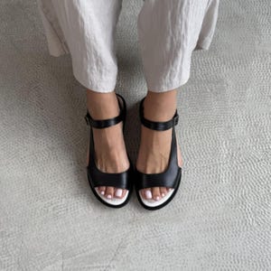 Puede incluir: Sandalias de cuero negro con suela blanca y correa en el tobillo. Las sandalias tienen un dise&ntilde;o simple y moderno con cierre de hebilla. El calzado se muestra desde arriba.