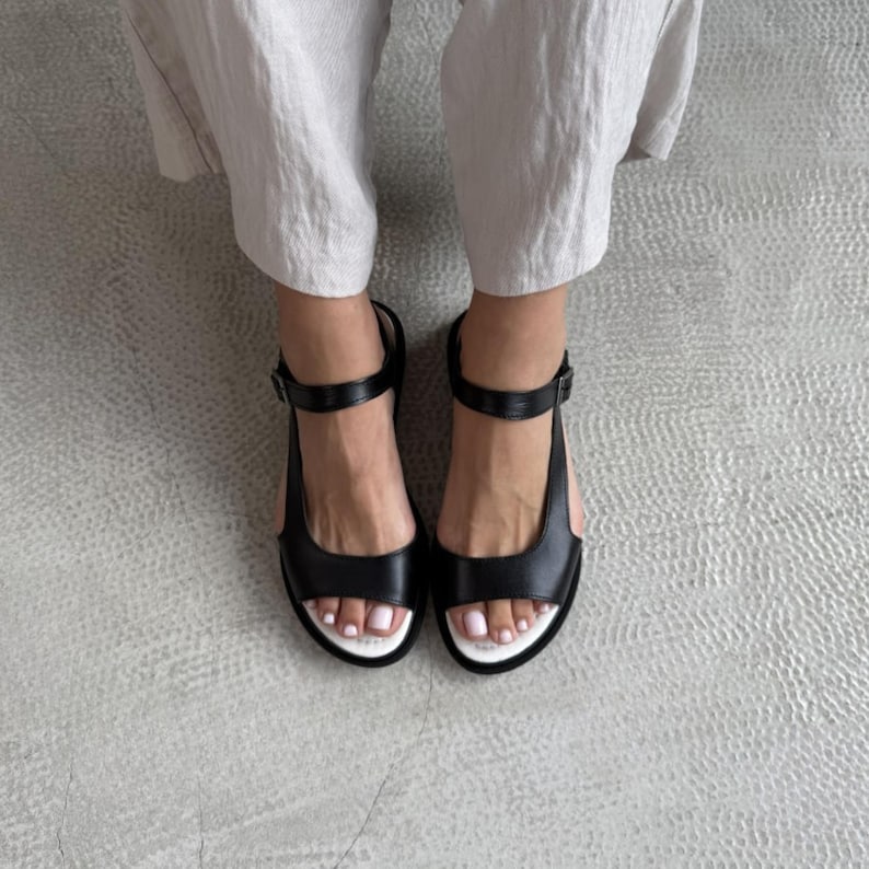 Puede incluir: Sandalias de cuero negro con suela blanca y correa en el tobillo. Las sandalias tienen un dise&ntilde;o simple y moderno con cierre de hebilla. El calzado se muestra desde arriba.