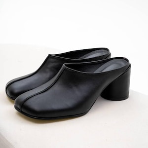 Puede incluir: Un par de mulas de cuero negro con punta redondeada y tacón de bloque. Los zapatos de espalda abierta tienen un acabado liso y mate y un detalle de costura en el centro de la pala. Los tacones miden unos 7,6 cm de alto.