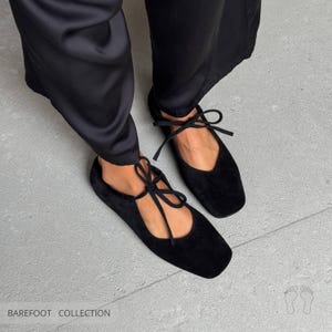 May include: Black suede ballet flats with square toes and delicate tie-up ankle straps. The flats are part of the Barefoot Collection. The shoes are shown with black satin pants on a gray concrete surface.