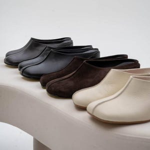 Puede incluir: Una colección de zapatos sin cordones de cuero y ante en varios colores, incluyendo negro, marrón oscuro y crema. Los zapatos están dispuestos en fila, mostrando sus diferentes texturas y estilos. El calzado se exhibe sobre una superficie blanca.