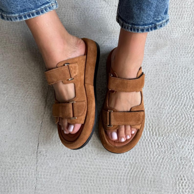 May include: Brown suede sandals with two adjustable straps and buckle closures. The sandals have a thick sole and a black base. The image shows the sandals being worn.