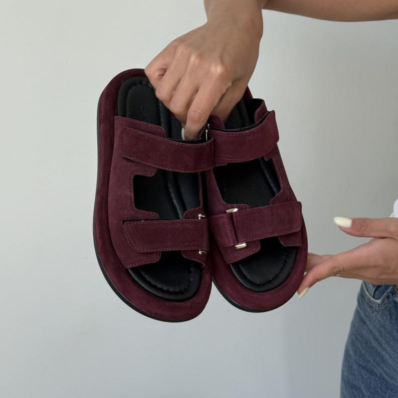 May include: A pair of burgundy suede sandals with adjustable straps and black soles. The sandals have a platform sole and are held up by a person's hands. The sandals are a stylish footwear option.