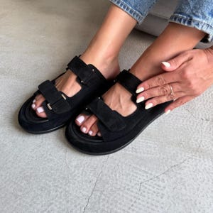 May include: Black suede sandals with adjustable hook-and-loop straps. The sandals have a thick sole and are worn by a person. The sandals are a casual style.