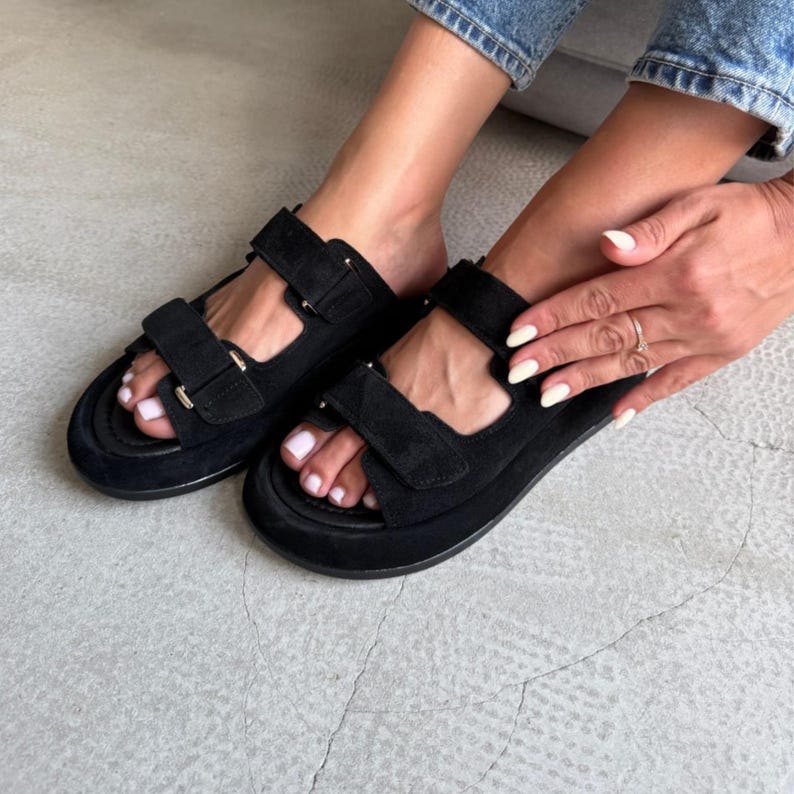 May include: Black suede sandals with adjustable hook-and-loop straps. The sandals have a thick sole and are worn by a person. The sandals are a casual style.