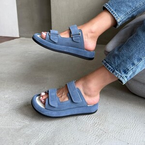 May include: Light blue suede sandals with two adjustable straps. The sandals have a thick black sole and a white footbed. The image shows the sandals being worn, with the feet and lower legs visible.