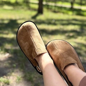 Puede incluir: Un par de zapatos sin cordones de ante marrón con suela negra y un pequeño detalle de hebilla. Los zapatos se muestran en un entorno al aire libre con un fondo verde borroso.