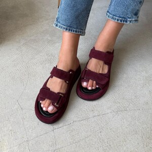 May include: Burgundy suede sandals with adjustable straps. The sandals have a thick black sole and are worn with blue jeans. The sandals are a slip-on style with two straps across the top of the foot.