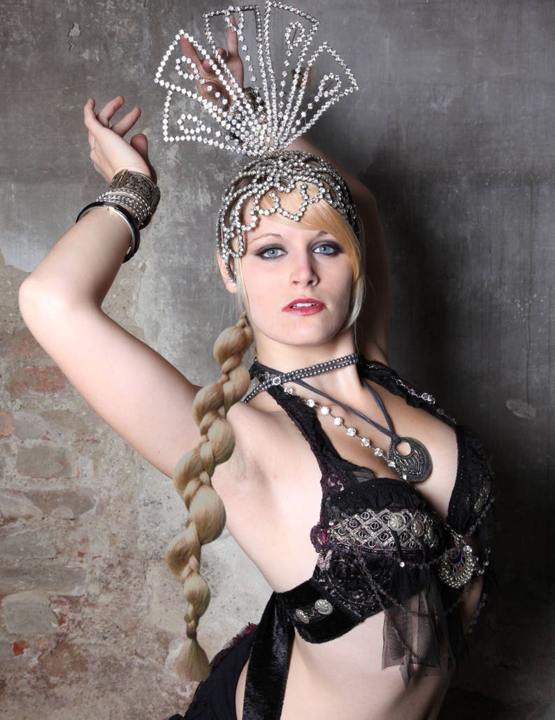 Op de afbeelding: Een vrouw draagt een zilveren en strass-hoofdtooi, een zwarte bh-top met zilveren versieringen en een lange vlecht. Ze poseert met haar arm omhoog.