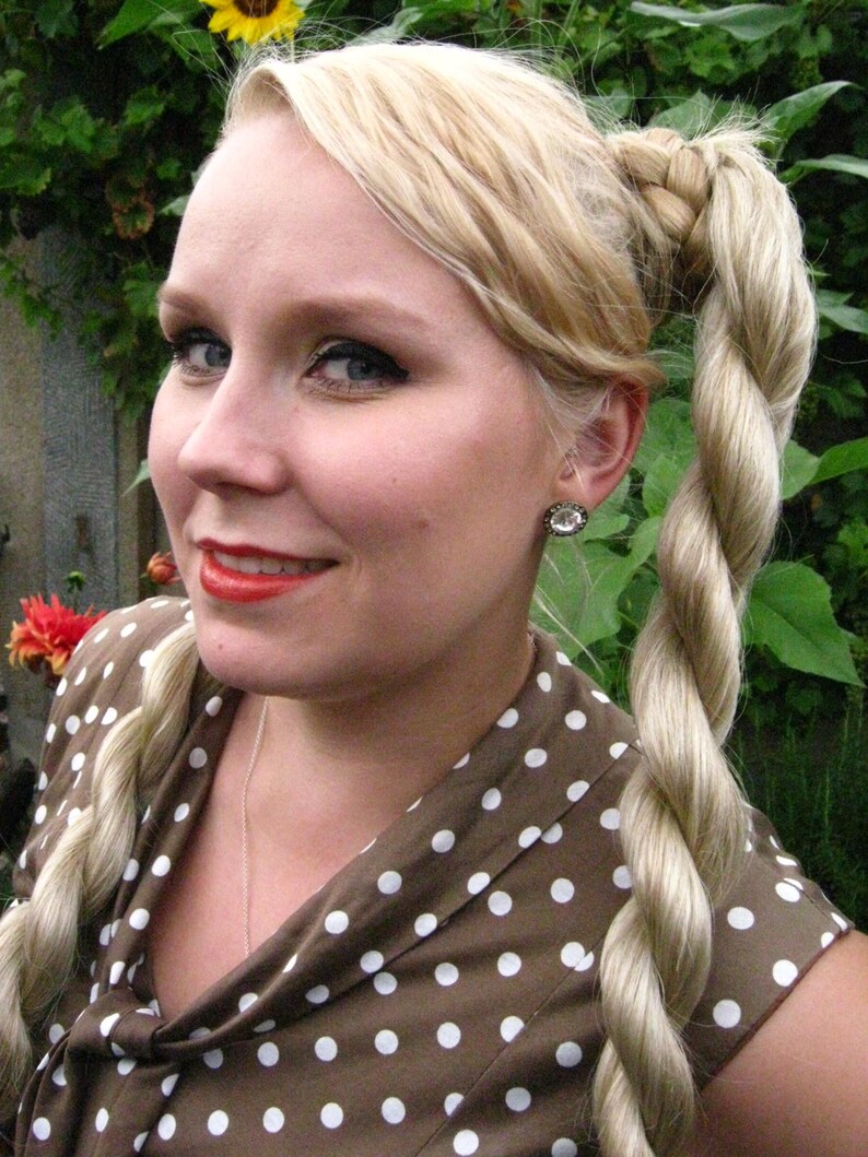 May include: A woman with blonde hair styled in a braided ponytail, wearing a brown and white polka dot dress. She is wearing a silver necklace and a small earring.