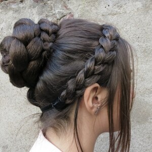 May include: A woman with long brown hair styled in a braided crown and a bun. The bun is secured with a black elastic band.