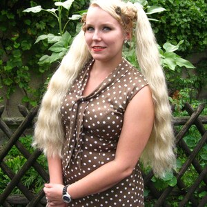May include: A woman with long blonde hair styled in pigtails with hair clips is wearing a brown dress with white polka dots. She is standing in front of a wooden fence with green plants.