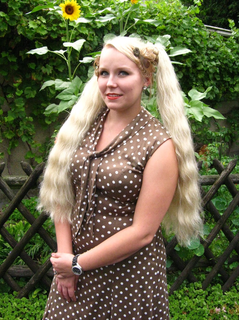May include: A woman with long blonde hair styled in pigtails with hair clips is wearing a brown dress with white polka dots. She is standing in front of a wooden fence with green plants.