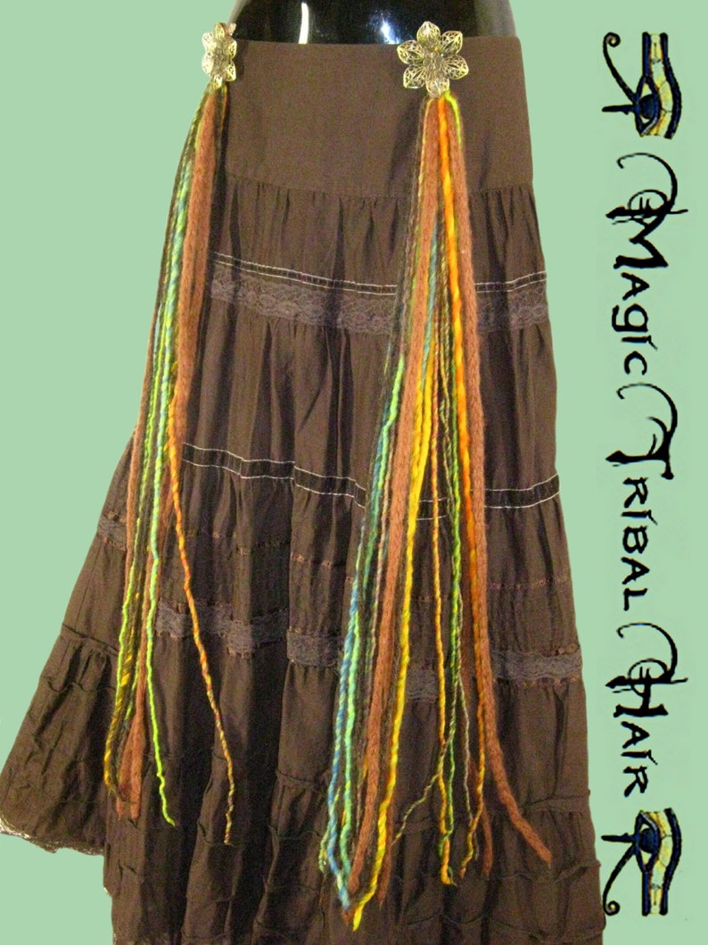 May include: A brown skirt with multiple tiers and lace trim. Two gold flower-shaped clips are attached to the skirt, each with colorful yarn strands hanging down.