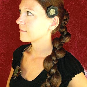 Op de afbeelding: Een vrouw met lang bruin haar dat in een dikke touw gevlochten is. Ze draagt een zwart topje met een kleine gouden en zwarte bloemspeld in haar haar.