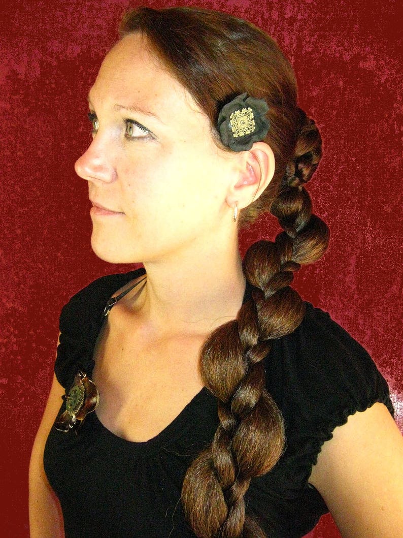 Op de afbeelding: Een vrouw met lang bruin haar dat in een dikke touw gevlochten is. Ze draagt een zwart topje met een kleine gouden en zwarte bloemspeld in haar haar.