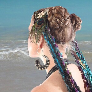 May include: A woman with long brown hair styled in two braids, wearing a peacock feather headband with turquoise and purple fabric strands hanging down her back. She is wearing large silver hoop earrings with intricate designs.