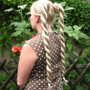 May include: A woman with long blonde hair styled in two thick braids, wearing a brown dress with white polka dots. She is standing in a garden with green foliage and sunflowers.
