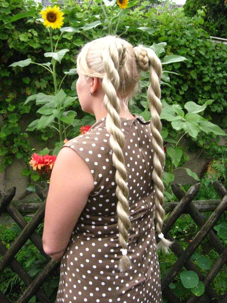 May include: A woman with long blonde hair styled in two thick braids, wearing a brown dress with white polka dots. She is standing in a garden with green foliage and sunflowers.