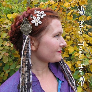 K&ouml;nnte beinhalten: Eine Frau mit roten Haaren tr&auml;gt ein lilafarbenes Hemd und ein Haarschmuck mit wei&szlig;en Blumen und einem Metallanh&auml;nger. Der Haarschmuck ist an einer langen, braunen, wei&szlig;en und grauen geflochtenen Schnur befestigt.