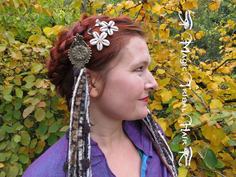 K&ouml;nnte beinhalten: Eine Frau mit roten Haaren tr&auml;gt ein lilafarbenes Hemd und ein Haarschmuck mit wei&szlig;en Blumen und einem Metallanh&auml;nger. Der Haarschmuck ist an einer langen, braunen, wei&szlig;en und grauen geflochtenen Schnur befestigt.
