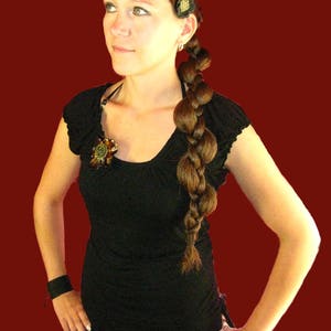 Op de afbeelding: Een vrouw met lang bruin haar dat in een vlecht is gevlochten, draagt een zwart topje met een bloemversiering en een bruine rok.