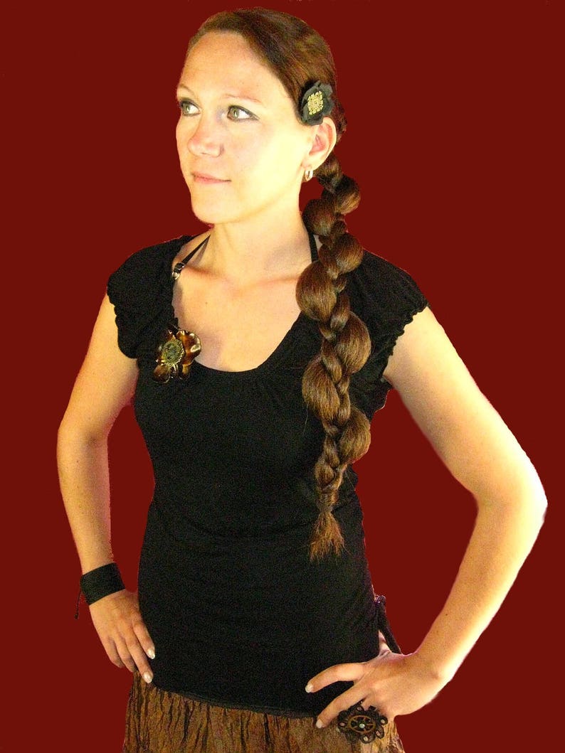 Op de afbeelding: Een vrouw met lang bruin haar dat in een vlecht is gevlochten, draagt een zwart topje met een bloemversiering en een bruine rok.