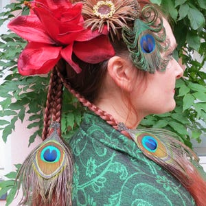 May include: A woman with red hair wearing a green shawl with a floral pattern. She has a large red flower and a brown flower with a gold center in her hair. She also has two peacock feathers in her hair and two more peacock feathers tied into her braids.