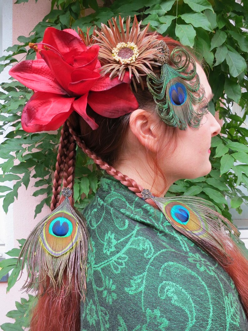 May include: A woman with red hair wearing a green shawl with a floral pattern. She has a large red flower and a brown flower with a gold center in her hair. She also has two peacock feathers in her hair and two more peacock feathers tied into her braids.