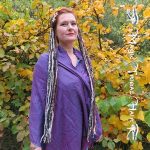 K&ouml;nnte beinhalten: Eine Frau mit langen, bunten Haarverl&auml;ngerungen in einem lila Shirt steht vor einem Busch mit gelben Bl&auml;ttern. Die Haarverl&auml;ngerungen sind aus Garn und mit wei&szlig;em Garn gebunden. Der Text "Magic Tribal Hair" ist auf der rechten Seite des Bildes sichtbar.
