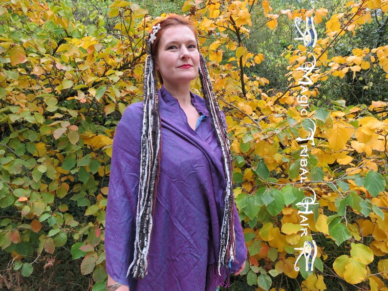 K&ouml;nnte beinhalten: Eine Frau mit langen, bunten Haarverl&auml;ngerungen in einem lila Shirt steht vor einem Busch mit gelben Bl&auml;ttern. Die Haarverl&auml;ngerungen sind aus Garn und mit wei&szlig;em Garn gebunden. Der Text "Magic Tribal Hair" ist auf der rechten Seite des Bildes sichtbar.