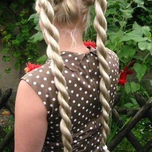May include: A woman with long blonde hair styled in two thick braids. She is wearing a brown and white polka dot dress.