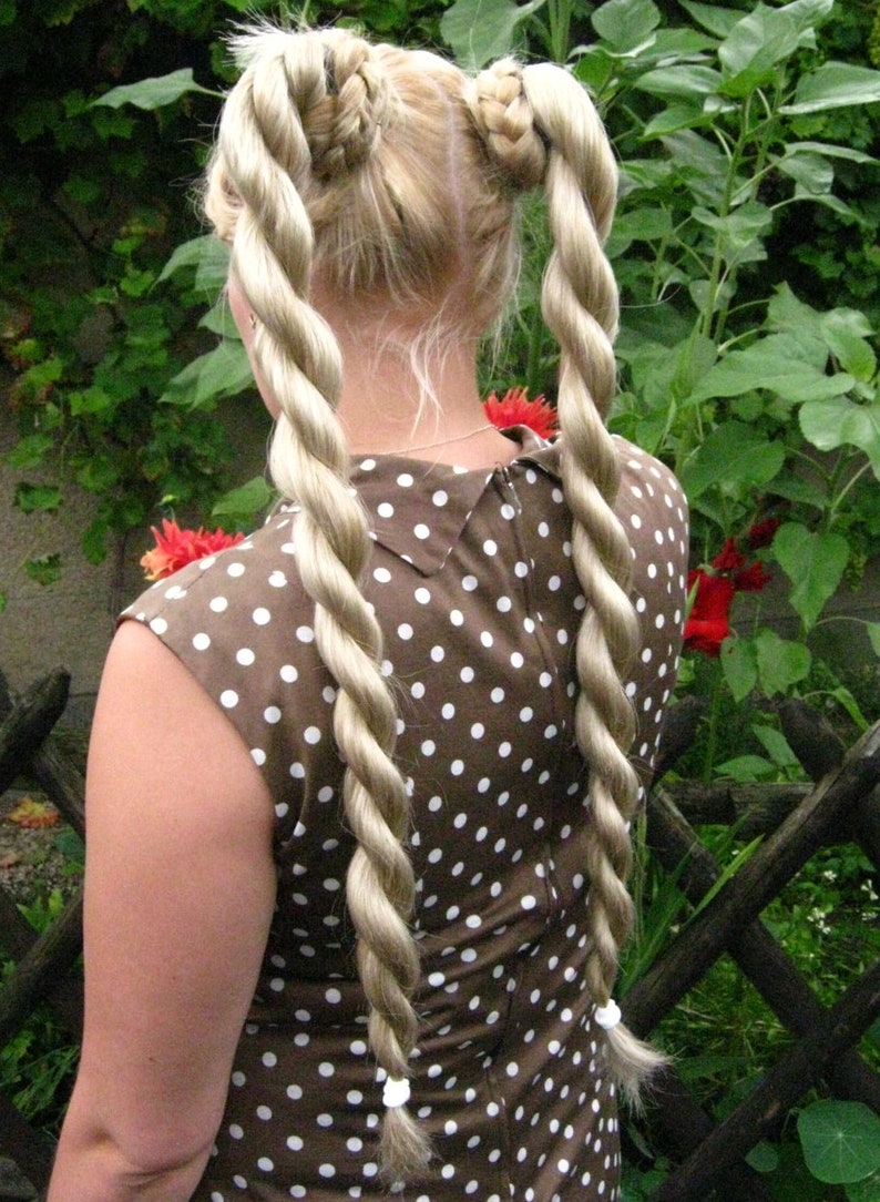 May include: A woman with long blonde hair styled in two thick braids. She is wearing a brown and white polka dot dress.