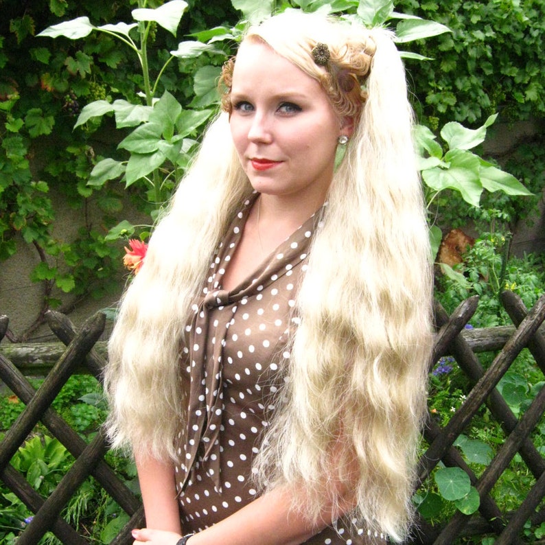 May include: A woman with long blonde hair styled in two pigtails with brown hair clips. She is wearing a brown and white polka dot dress with a bow at the neck. She is standing in front of a wooden fence and green foliage.
